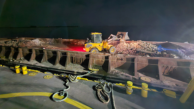 A front-end loader moves cobble around on a barge at night. The scene is lit by work lights and the taillights of the loader bathe the barge in red. Multiple large cleats are in the foreground of the photo on the edge of the barge the photographer is standing on.